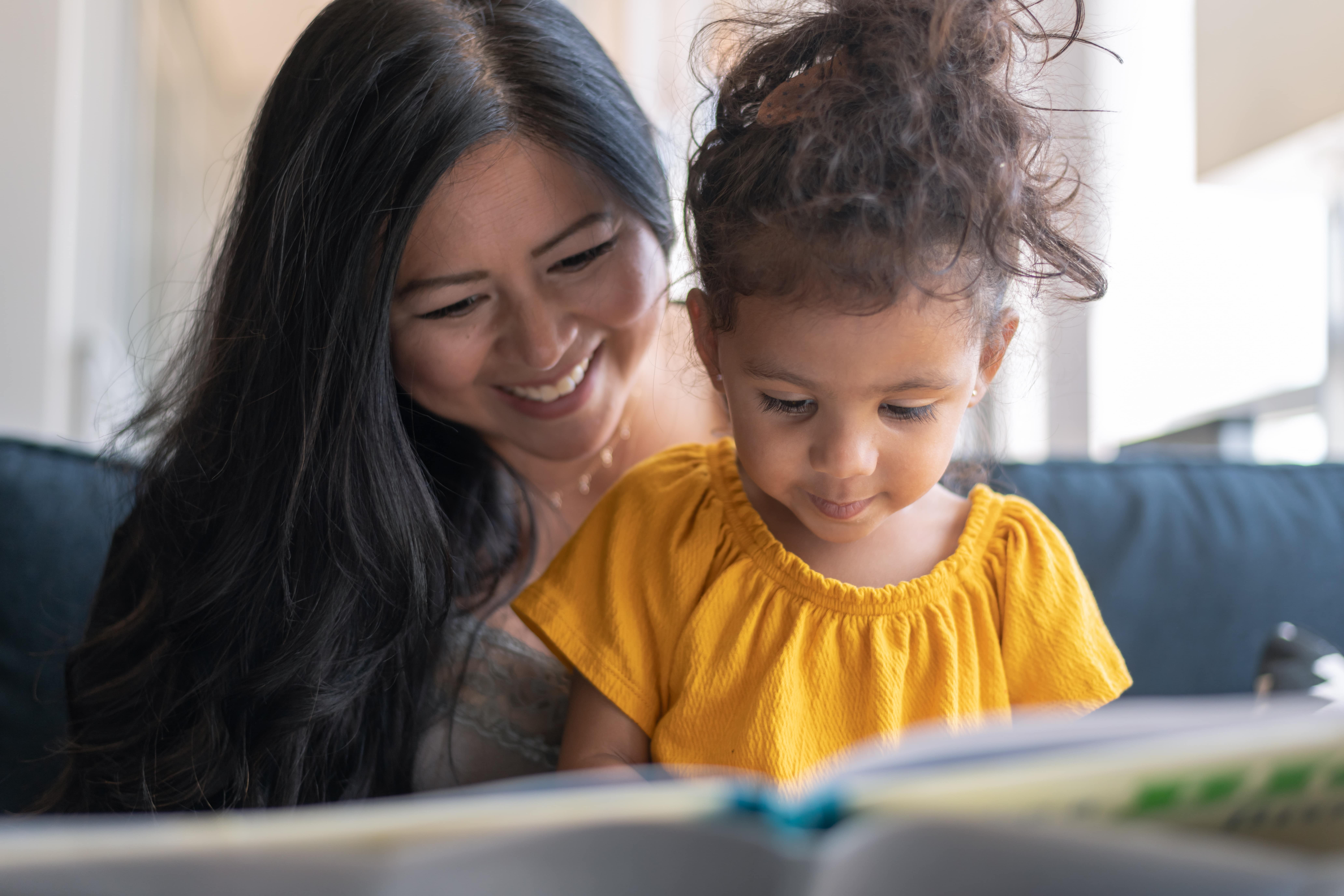 A loving mother of Asian descent sits on the couch at home and reads a storybook to her preschool age daughter. The child is sitting on her mother's lap and looking at the book intently.
